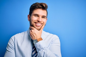Young blond businessman with beard and blue eyes wearing elegant shirt and tie standing looking confident at the camera smiling with crossed arms and hand raised on chin. Thinking positive.