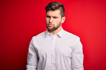 Young blond businessman with beard and blue eyes wearing elegant shirt over red background skeptic and nervous, frowning upset because of problem. Negative person.