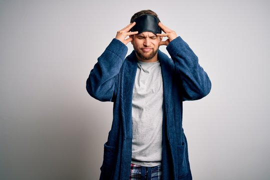 Young blond man with beard and blue eyes wearing sleep mask and pajama suffering from headache desperate and stressed because pain and migraine. Hands on head.