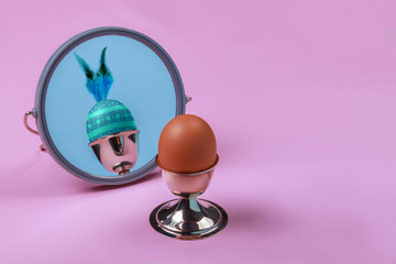 Happy Easter concept. Brown egg in silver cup reflected in a mirror as colored egg with feathers. Pink and blue background. Free copy space