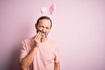 Middle age hoary man wearing bunny ears standing over isolated pink background touching mouth with hand with painful expression because of toothache or dental illness on teeth. Dentist