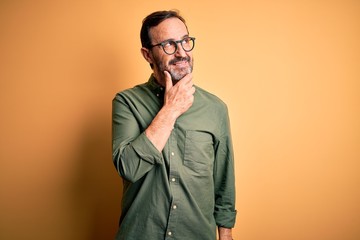 Fototapeta premium Middle age hoary man wearing casual green shirt and glasses over isolated yellow background with hand on chin thinking about question, pensive expression. Smiling with thoughtful face. Doubt concept.