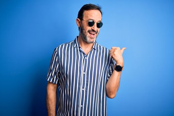 Middle age hoary man wearing striped shirt and sunglasses over isolated blue background smiling with happy face looking and pointing to the side with thumb up.