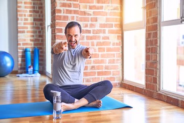 Naklejka premium Middle age handsome sportman sitting on mat doing stretching yoga exercise at gym pointing to you and the camera with fingers, smiling positive and cheerful