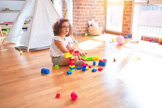 Beautiful toddler wearing glasses and unicorn diadem sitting playing with building blocks at kindergarten