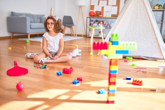 Beautiful toddler wearing glasses and unicorn diadem sitting on the floor playing with building blocks smiling at kindergarten