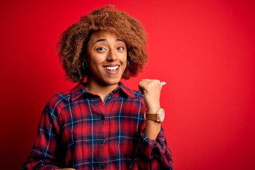 Young beautiful African American afro woman with curly hair wearing casual shirt smiling with happy face looking and pointing to the side with thumb up.