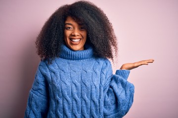 Young beautiful african american woman with afro hair wearing winter sweater over pink background smiling cheerful presenting and pointing with palm of hand looking at the camera.