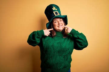 Young beautiful plus size woman wearing green hat with clover celebrating saint patricks day smiling cheerful showing and pointing with fingers teeth and mouth. Dental health concept.