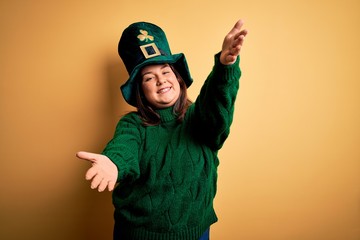 Young beautiful plus size woman wearing green hat with clover celebrating saint patricks day looking at the camera smiling with open arms for hug. Cheerful expression embracing happiness.