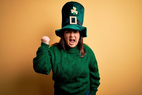 Young beautiful plus size woman wearing green hat with clover celebrating saint patricks day angry and mad raising fist frustrated and furious while shouting with anger. Rage and aggressive concept.