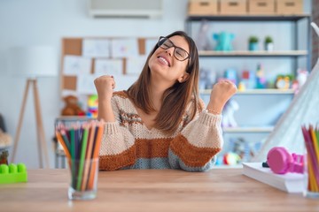 Young beautiful teacher woman wearing sweater and glasses sitting on desk at kindergarten...