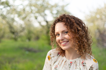 Close up portrait of young smiling attractive woman with curly hair in green flowering spring park. Pure emotions.