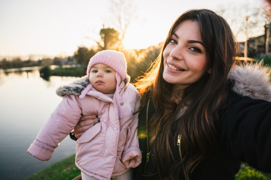 Mom takes a selfie at the park with her little daughter in her arms at sunset with a lake behind them - Moment of intimacy of the mother with the child