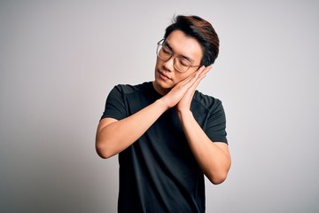 Young handsome chinese man wearing black t-shirt and glasses over white background sleeping tired dreaming and posing with hands together while smiling with closed eyes.