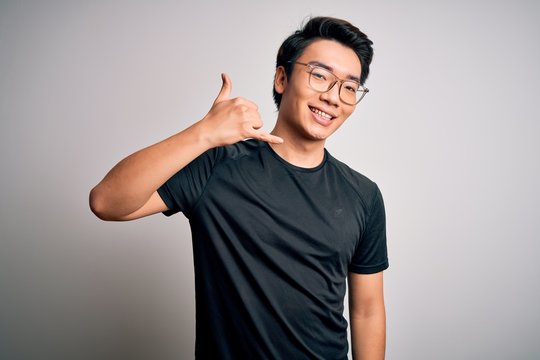 Young handsome chinese man wearing black t-shirt and glasses over white background smiling doing phone gesture with hand and fingers like talking on the telephone. Communicating concepts.