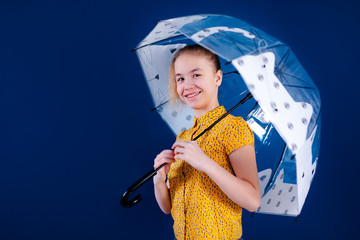 Happy teenage girl in yellow shirt with transparent umbrella posing on blue studio background.