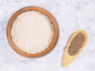 Indian long grain Basmati rice in brown wooden bowl and cumin (jeera) spoon on white concrete background