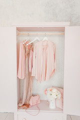 wooden pink wardrobe for women’s clothes, open doors, decor, mirror, bag, hangers, silk dresses in a bright room, concept, flowers delicate pastel colors