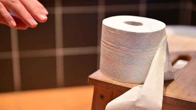 Close-up image of the toilet paper.In the blurred background you can recognize the bare legs of a Caucasian man as he defecates.The hands drum the fingers.The right hand tries to grab the toilet paper