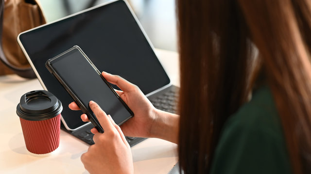 Cropped image of long hair woman holding smartphone in her hands in front a computer tablet next to takeaway coffee cup at the modern working table. Woman lifestyle, Woman relaxation concept.
