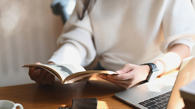 Cropped image of young beautiful woman holding/reading a books in her hands in front a computer laptop, coffee cup and smartphone at the modern wooden table with comfortable living room as background.