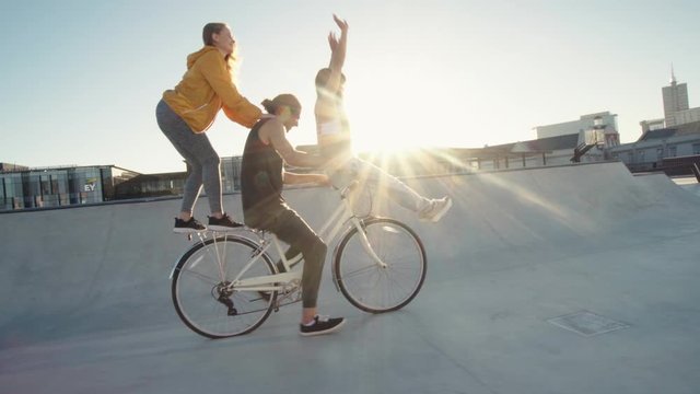 Man on bicycle with a female friend sitting on the handlebar and another standing in back enjoying and raising their hands.