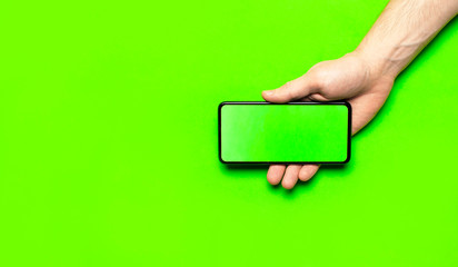 Male hands hold a modern black smartphone with green blank screen on neon green background flat lay top view. Modern technology, phone, gadget in hands, touch screen, template for your design. Mockup