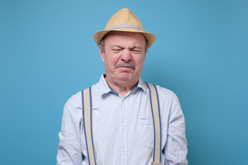 Senior man in summer hat with disgusted expression repulsing something. Disgust concept. Human emotions, facial expression concept. Studio shot blue wall.