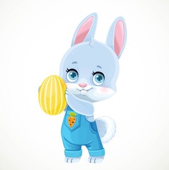 Cute little bunny in denim overalls holds yellow Easter painted egg in paws isolated on white background