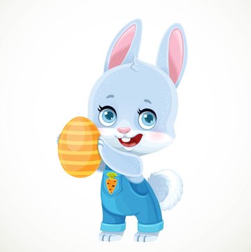 Cute little bunny in denim overalls holds Easter painted egg in paws isolated on white background