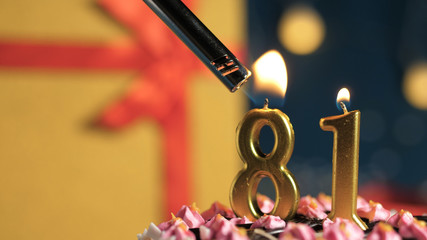 Birthday cake number 81 golden candles burning by lighter, background gift yellow box tied up with red ribbon. Close-up