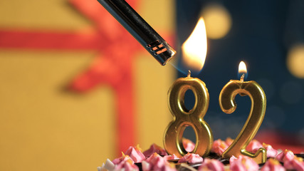 Birthday cake number 82 golden candles burning by lighter, background gift yellow box tied up with red ribbon. Close-up