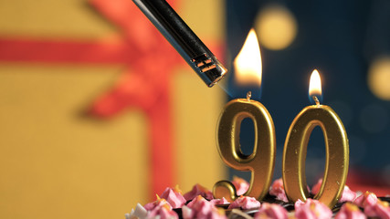Birthday cake number 90 golden candles burning by lighter, background gift yellow box tied up with...