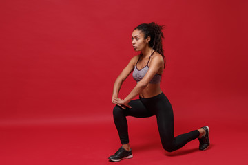 Fototapeta na wymiar Confident young african american sports fitness woman in sportswear posing working out isolated on red wall background. Sport exercises healthy lifestyle concept. Stretching her legs, doing lunges.