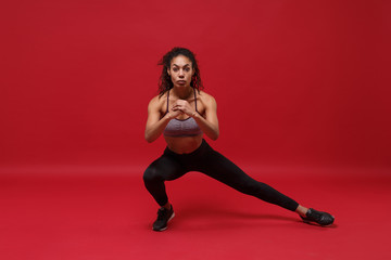 Fototapeta na wymiar Beautiful young african american sports fitness woman in sportswear posing working out isolated on red wall background studio portrait. Sport exercises healthy lifestyle concept. Stretching her legs.