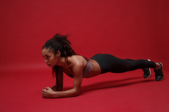 Strong beautiful young african american sports fitness woman in sportswear working out isolated on red background studio portrait. Sport exercises healthy lifestyle concept. Doing plank exercises