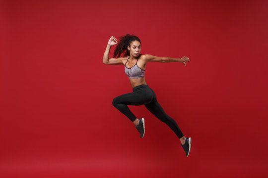 Side view of young african american fitness woman in sportswear posing working out isolated on red wall background studio portrait. Sport exercises healthy lifestyle concept. Jumping like running.