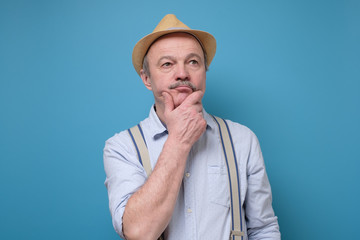 Senior man in summer hat thinking and rubbing a chin trying to make a right solution. Studio shot on blue wall