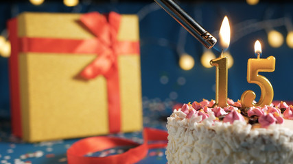 White birthday cake number 15 golden candles burning by lighter, blue background with lights and...