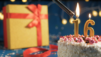 White birthday cake number 16 golden candles burning by lighter, blue background with lights and...