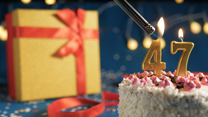 White birthday cake number 47 golden candles burning by lighter, blue background with lights and gift yellow box tied up with red ribbon. Close-up