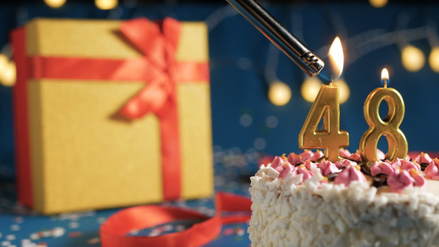White birthday cake number 48 golden candles burning by lighter, blue background with lights and gift yellow box tied up with red ribbon. Close-up