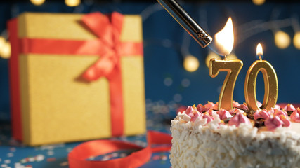 White birthday cake number 70 golden candles burning by lighter, blue background with lights and...