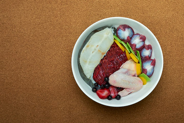 Natural raw organic dog food in bowl on brown floor background. Meat fish chicken vegetables fruits