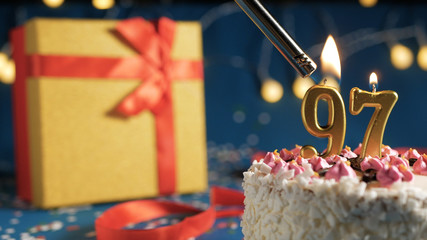 White birthday cake number 97 golden candles burning by lighter, blue background with lights and...