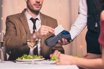 cropped view of elegant man paying with credit card with terminal while sitting in restaurant near girlfriend