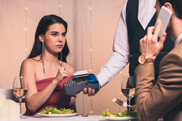 displeased girl paying with credit card with terminal while sitting at served table and looking at...