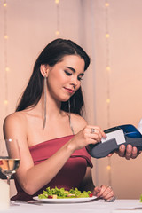 Fototapeta na wymiar attractive, elegant girl sitting at served table in restaurant and paying by credit card with terminal