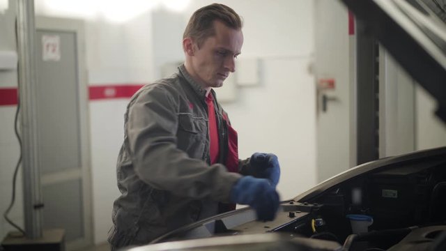 Professional Caucasian auto mechanic checking engine oil. Serious young man performing car inspection in repair shop. Auto insurance, tuning, automotive industry.